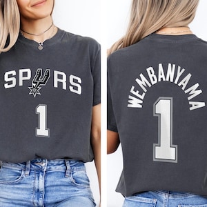 Op de afbeelding: Donkergrijs t-shirt met het woord "SPURS" in witte blokletters en een teamlogo. Het nummer "1" staat onder de tekst. De achterkant van het shirt toont "WEMBANYAMA" boven het nummer "1".