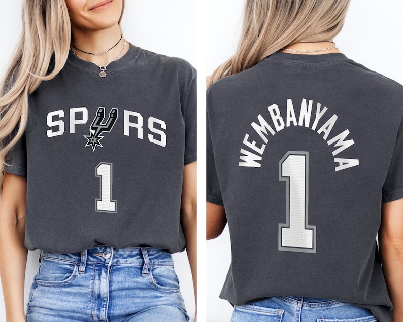 Op de afbeelding: Donkergrijs t-shirt met het woord "SPURS" in witte blokletters en een teamlogo. Het nummer "1" staat onder de tekst. De achterkant van het shirt toont "WEMBANYAMA" boven het nummer "1".