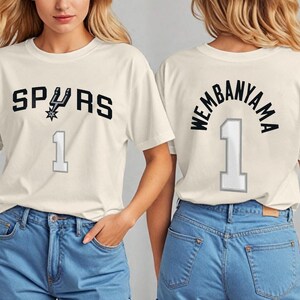 Op de afbeelding: Cr&egrave;mekleurig T-shirt met het woord "SPURS" in zwarte blokletters, een teamlogo en het nummer "1" in grijs. De achterkant van het shirt toont "WEMBANYAMA" boven het nummer "1".