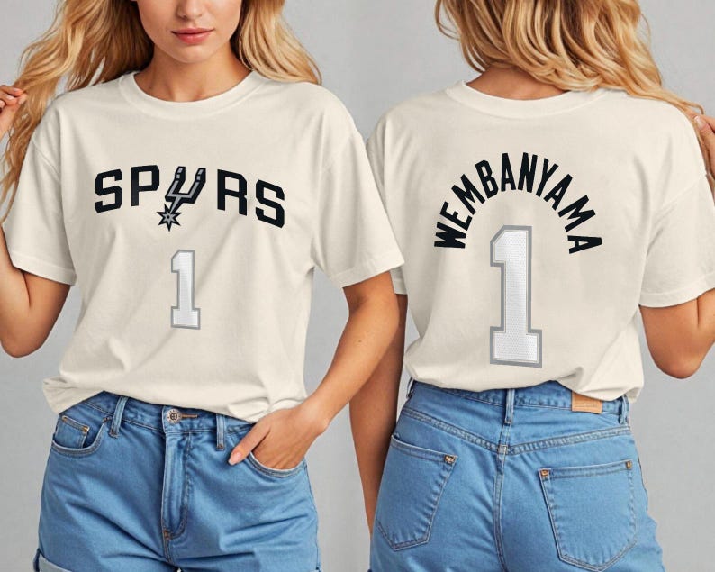 Op de afbeelding: Cr&egrave;mekleurig T-shirt met het woord "SPURS" in zwarte blokletters, een teamlogo en het nummer "1" in grijs. De achterkant van het shirt toont "WEMBANYAMA" boven het nummer "1".
