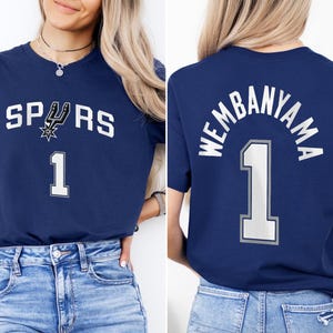 Op de afbeelding: Marineblauw T-shirt met het woord "SPURS" en het nummer "1" in het wit. Het Spurs-logo is ook afgebeeld. De achterkant van het shirt toont "WEMBANYAMA" boven het nummer "1".