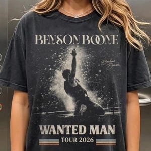 May include: A dark gray t-shirt featuring the text "BENSON BOONE" above a graphic of a person with an arm raised. Below the graphic are the words "WANTED MAN TOUR 2026" in a retro font. The shirt has a vintage, worn look.