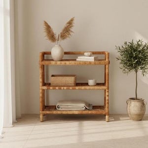 May include: A three-tiered light brown rattan shelving unit featuring a woven texture. The top shelf holds a white vase with dried pampas grass and stacked books. The middle shelf contains a woven basket and a small white candle.