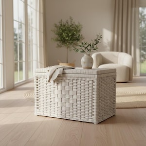 May include: A rectangular white woven storage chest with a flat lid. A folded light-coloured fabric cloth rests on top of the lid. The chest features a structured, grid-like weave pattern and sits on a light-coloured wooden floor surface.