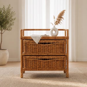 May include: A light brown rattan storage unit featuring two rectangular woven drawers stacked vertically. The top surface holds a white ceramic donut-shaped vase containing a dried pampas grass stem and a folded light beige cloth napkin.