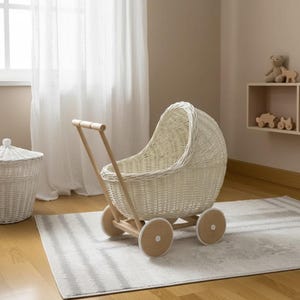 May include: A white wicker doll stroller with a wooden handle and wheels. The stroller has a rounded shape and is sitting on a gray and white striped rug. The wheels are made of wood and have a light brown color.
