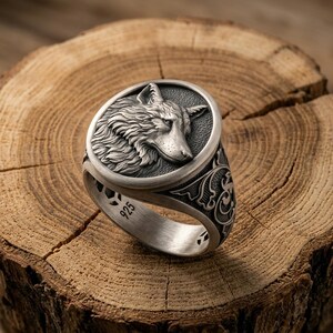 May include: A silver ring featuring a circular face with a detailed relief of a wolf head in profile. The band displays intricate scrollwork patterns on the sides and is stamped with the number 925 on the inner surface, indicating sterling silver.