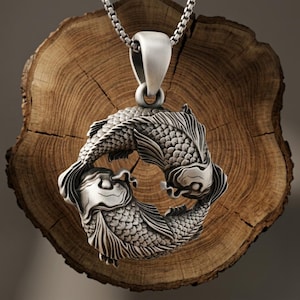 Peut inclure: Un pendentif en argent représentant deux poissons koï nageant en formation circulaire, créant une forme de yin-yang. Le pendentif présente une texture d'écailles détaillée et est suspendu à une chaîne en argent, avec une finition métallique oxydée.