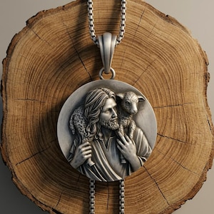 Puede incluir: Un colgante circular de tono plateado que presenta una escultura en relieve de una figura barbuda con cabello largo que lleva un pequeño cordero sobre sus hombros. El colgante está suspendido de una cadena de plata y tiene un acabado metálico.