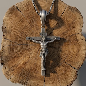 Puede incluir: Un colgante de crucifijo de metal plateado que presenta una figura detallada de Jesús en una cruz con las letras I.N.R.I. inscritas sobre la figura. El colgante está unido a un collar de cadena de caja plateada con un acabado pulido.