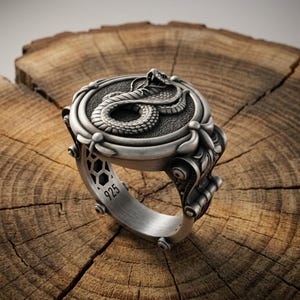 Puede incluir: Un anillo de plata de ley con una cara circular que presenta un relieve de cobra sobre un fondo texturizado. La banda muestra detalles intrincados de volutas y un sello distintivo 925 claramente visible en el interior del metal.