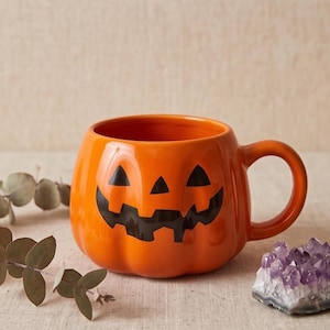 Peut inclure: Une tasse en céramique orange vif en forme de citrouille avec une finition brillante. La tasse présente un visage de citrouille d'Halloween peint en noir avec des yeux triangulaires et un sourire dentelé. Elle a un corps arrondi et une anse.