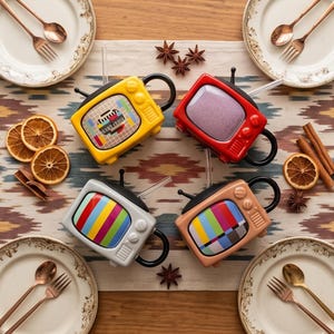 Può includere: Quattro tazze a forma di TV retrò in giallo, rosso, grigio e beige, ciascuna con un manico e un'antenna neri. Gli schermi mostrano modelli di test colorati. Le tazze sono disposte su una tovaglia fantasia con piatti, posate e spezie.