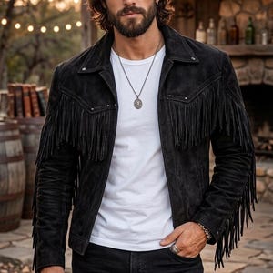 Peut inclure: Veste en daim noire avec des franges, portée sur un t-shirt blanc et un jean noir. La veste a un style western classique, associée à un chapeau de cowboy noir. Un collier et des bagues en argent complètent le look.