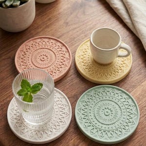 May include: A set of four circular coasters in pastel pink, yellow, white, and sage green, each featuring a raised geometric mandala pattern. One coaster holds a small white ceramic cup, another holds a clear ribbed glass with mint leaves.