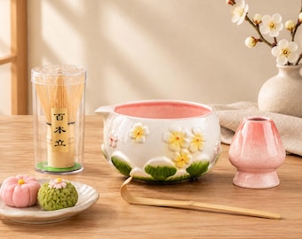 Könnte beinhalten: Ein Matcha-Teeset bestehend aus einem Bambusbesen in einer transparenten Kunststoffhülle mit japanischem Text, einer weißen Keramikschale mit gelben Blüten und grünen Blättern, einem rosa Besenhalter, einem Bambuslöffel und zwei kleinen Süßigkeiten.