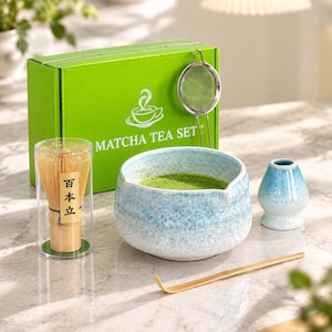 May include: A matcha tea set including a green box labeled MATCHA TEA SET, a metal strainer, a bamboo whisk in a clear case with Japanese text, a blue and white speckled ceramic bowl containing green tea, and a bamboo scoop.