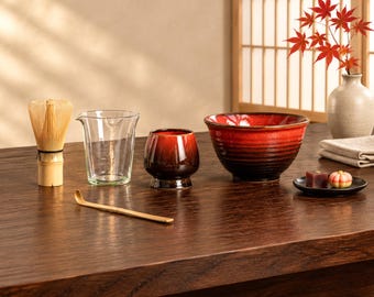 Puede incluir: Un batidor de matcha de bambú, una jarra de cristal, una taza pequeña de cerámica roja y negra, un cuenco grande de cerámica roja y negra, una cuchara de bambú y un plato pequeño con dos dulces. Un jarrón con hojas de arce rojas.