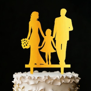 Puede incluir: Un adorno dorado para pastel con la silueta de una familia. La novia sostiene un ramo, el niño está en el medio y el novio a la derecha. El adorno está colocado sobre un pastel blanco con glaseado decorativo.