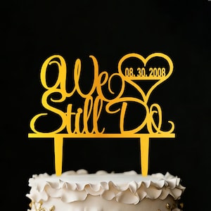 Può includere: Un cake topper dorato con le parole "We Still Do" in elegante calligrafia. La data "08.30.2008" è all'interno di un cuore. Il topper è posto su una torta bianca con glassa increspata, perfetto per una celebrazione dell'anniversario di matrimonio.