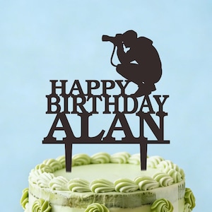 May include: A brown cake topper with the words "HAPPY BIRTHDAY ALAN" and a silhouette of a photographer taking a photo. The topper is placed on a cake with green frosting and decorations.