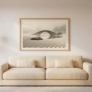 Op de afbeelding: Een ingelijste sepia-afdruk van een stenen brug die weerspiegeld wordt in het water, boven een beige bank met kussens. Het kunstwerk wordt verlicht door twee hanglampen. Het beeld toont een sereen, minimalistisch interieur.