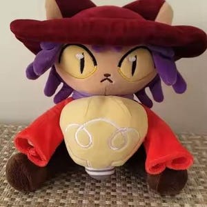 May include: A plush toy of a cat-like character with a maroon hat, purple hair, and a red jacket. The character has large yellow eyes and a yellow and white object in its hands. The toy is sitting on a textured surface.