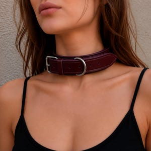 May include: A deep burgundy leather choker collar featuring a silver-toned metal buckle and a matching D-ring attachment. The collar has visible stitching along the edges and is secured around the neck with a standard pin-style buckle closure.