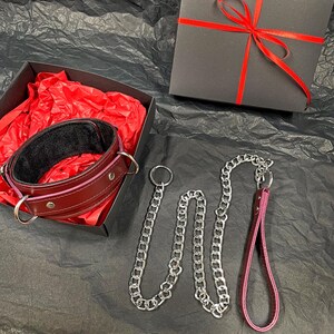 May include: A dark red leather collar with silver-toned metal rings and studs, featuring a black interior lining. The collar is placed inside a black gift box with red tissue paper. A matching dark red leather leash with a silver chain is included.