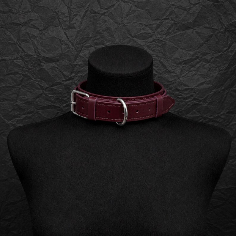 May include: A deep burgundy leather choker collar featuring a silver-toned metal buckle, a matching silver-toned D-ring, and multiple adjustment holes. The collar is constructed with visible stitching along the edges and a pointed strap end.