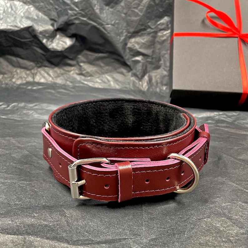 May include: A deep burgundy leather collar featuring a black interior lining, a silver-toned metal buckle, and a matching D-ring. The collar is constructed with visible stitching along the edges and includes a series of adjustment holes.
