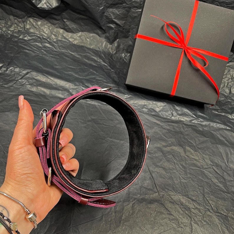 May include: A deep purple leather collar featuring a black interior lining, silver-toned metal hardware, and a buckle closure. The collar is presented alongside a square black gift box tied with a thin red ribbon, creating a complete set.