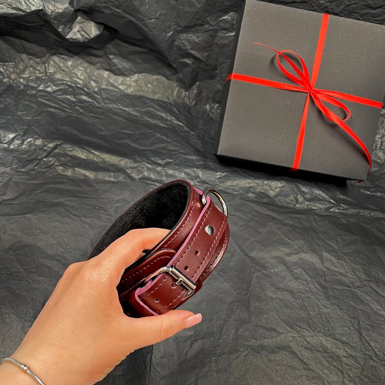 May include: A dark burgundy leather collar featuring a black interior lining, a silver-toned metal buckle, a D-ring, and a matching black gift box tied with a thin red ribbon. The collar has visible stitching along the edges and a metal stud.