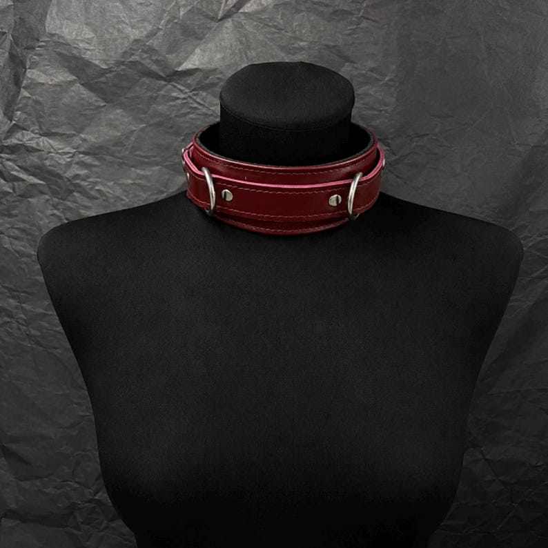 May include: A deep red leather choker collar featuring a wide band design with silver-toned metal hardware. The collar includes two circular rings positioned on the sides, secured by silver rivets along the length of the smooth, dark red strap.