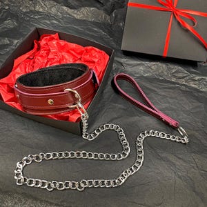May include: A deep burgundy leather collar with a black interior lining, silver-toned metal rivets, and a D-ring. A matching burgundy leather leash with a silver-toned metal chain and a loop handle is attached to the collar, presented in a gift box.