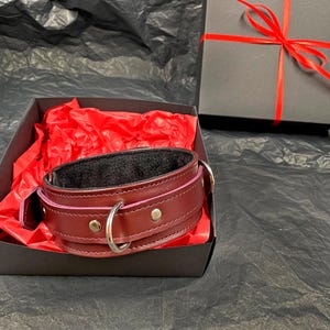 May include: A dark burgundy leather collar featuring a black interior lining, silver-toned metal hardware, and a central D-ring. The collar is presented inside an open black gift box lined with red tissue paper, with the box lid resting nearby.