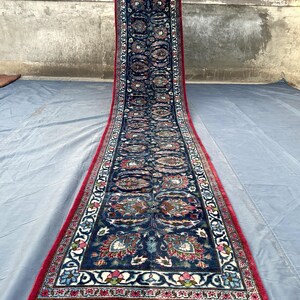 Könnte beinhalten: Ein langer, schmaler Läufer mit einem tiefblauen Mittelfeld, das mit sich wiederholenden Blumen- und Paisleuchtenden Blumen- und Paisleuchtenden Blumen- und Paisley-Motiven in Rot, Weiß und Hellblau verziert ist. Der Teppich ist von einem leuchtend roten Rand umgeben.