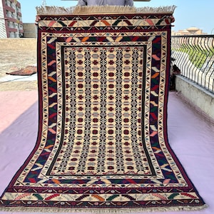 Peut inclure: Un tapis rectangulaire présentant un champ central de couleur crème avec des motifs floraux géométriques répétitifs en rouge foncé, bleu et vert. La bordure affiche un motif complexe de zigzags et de losanges colorés avec une frange.