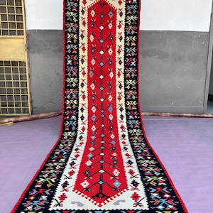 Puede incluir: Una alfombra larga y rectangular con un campo central rojo vibrante. La alfombra presenta un patrón geométrico repetitivo en negro, blanco y varios colores a lo largo de los bordes y un diseño central. La alfombra tiene un borde con flecos.