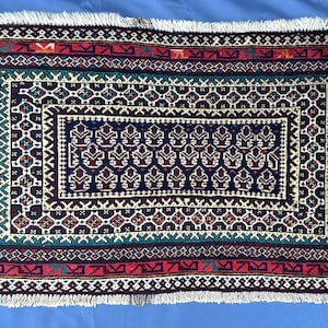 May include: A rectangular woven rug featuring a dark blue central field with repeating stylised floral motifs. The rug is framed by multiple decorative borders in shades of red, teal, and white, finished with white fringe on the top and bottom edges.