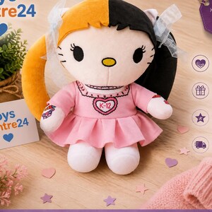 May include: A plush doll with a split orange and black hairstyle, wearing a pink dress with a heart-shaped pendant. The doll has white legs and arms, and is on a wooden surface. The doll is a collectible item.