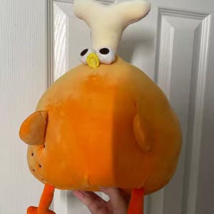 May include: A plush toy shaped like a fried chicken leg. The toy is primarily orange with a white bone-shaped piece on top. It has small black dots, orange legs, and cartoonish eyes.