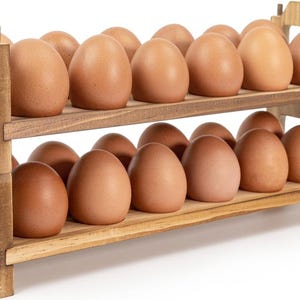May include: A two-tiered wooden egg holder filled with brown eggs. The holder is made of light-colored wood and has a simple, rustic design. Each tier holds a row of eggs, showcasing a farm-fresh aesthetic.