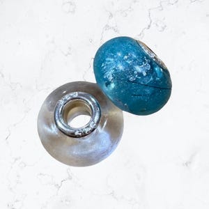 May include: Two decorative beads. One is clear with a silver-coloured metal ring, and the other is a blue-green colour with a silver-coloured design. The beads are likely for jewellery making or other craft projects.