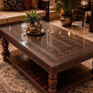 Op de afbeelding: Een rechthoekige houten salontafel met ingewikkelde, met de hand gesneden bloemmotieven op het oppervlak, afgedekt met een heldere glasplaat. De tafel rust op vier dikke, gedraaide poten en heeft een onderste plank voor opslag.