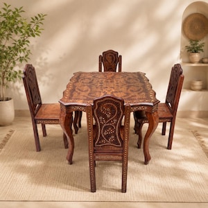 Op de afbeelding: Een houten eettafel en vier stoelen set. De tafel en stoelen zijn donkerbruin met ingewikkelde gesneden ontwerpen en decoratieve accenten. De tafel heeft gebogen poten en een gedetailleerd tafelblad. De stoelen hebben sierlijke rugleuningen.