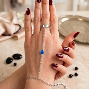 Könnte beinhalten: Ein silbernes Handkettenarmband mit einem Ring, einem blauen kreisförmigen Stein und einer Kette, die den Ring mit dem Armband verbindet. Die Hand auf dem Bild hat dunkelroten Nagellack. Der Hintergrund ist unscharf.