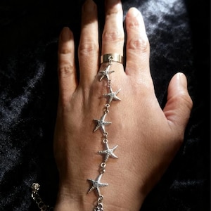 Puede incluir: Una cadena de mano plateada con un anillo y dijes de estrella de mar conectados por delicadas cadenas. La cadena de mano se usa en una mano, con el anillo en el dedo índice y la cadena que se extiende hasta la muñeca.