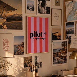 Könnte beinhalten: Ein gerahmter Wandkunstdruck mit vertikalen rosa und roten Streifen, dem schwarzen Text "pilot not passenger" und einem kleinen Flugzeugsymbol. Der Druck hängt an einer Wand, umgeben von verschiedenen anderen gerahmten Fotos und Reisebildern.