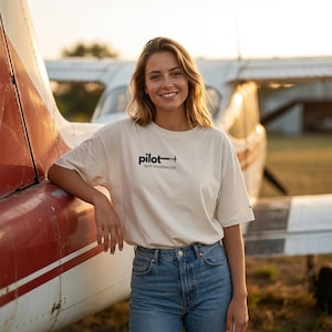 Könnte beinhalten: Cremefarbenes T-Shirt mit dem schwarzen Aufdruck "pilot - not passenger". Das Shirt wird mit einer blauen Jeans kombiniert. Im Hintergrund ist ein rot-weißes Flugzeug zu sehen.